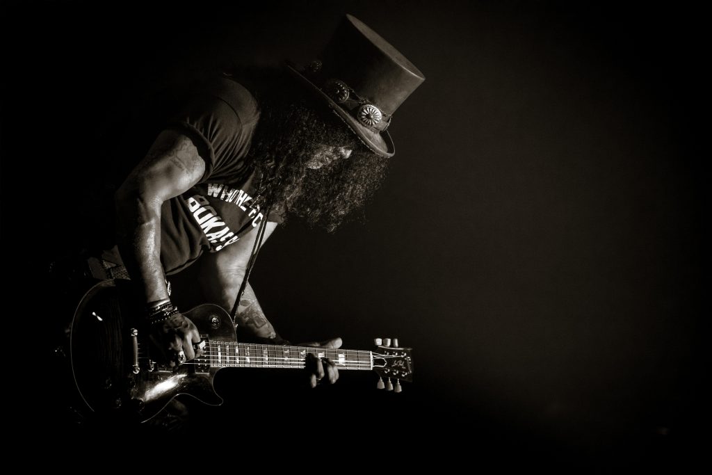 Slash in top hat playing guitar in a single beam of light