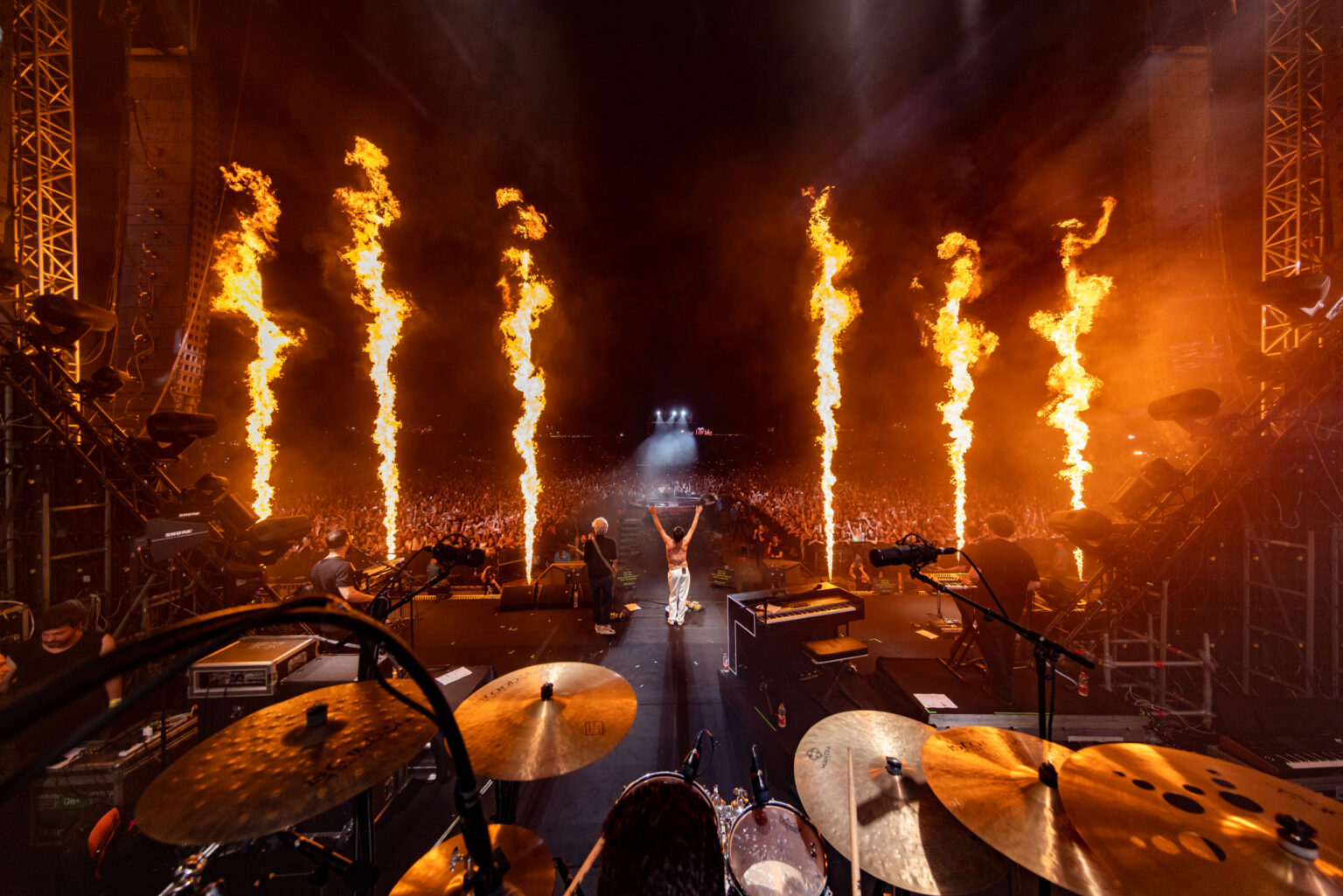 Arms-spread frontman lit by flame columns in front of a festival crowd, shot from behind the drum kit